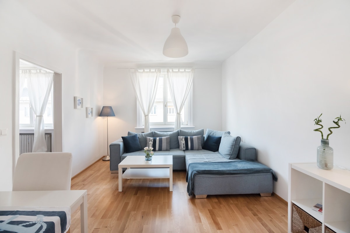 A bright living room features a spacious blue sectional sofa and a white coffee table. Light-colored wooden floors provide warmth to the space, while large windows allow natural light to fill the room. Decorative plants add a touch of greenery to the minimalist decor.
