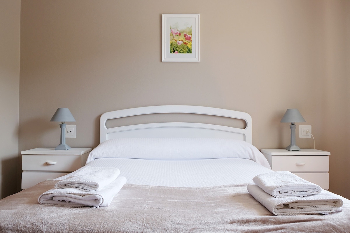 A light-colored bedroom features a spacious bed adorned with fresh linens. Two bedside tables, each with a small lamp, flank the bed. Above the headboard, a framed artwork depicting flowers adds a subtle pop of color to the neutral walls.