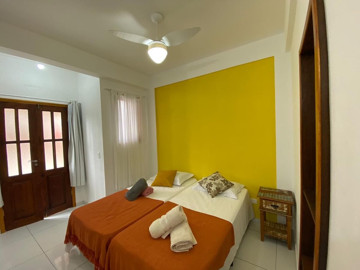 A bright room features two spacious beds adorned with light blankets. A vibrant yellow accent wall enhances the space. A ceiling fan is present, and natural light enters through a window with curtains. A wooden cabinet stands beside the beds, contributing to the room's functionality.