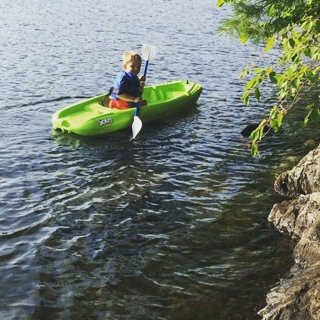 A brightly colored kayak sits on the calm water, with a young child paddling. The shoreline features lush greenery, and a prominent rock formation adds texture to the scene.