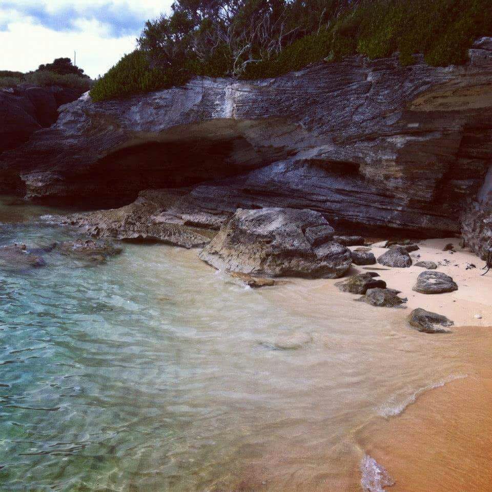 A serene beach scene features gentle waves lapping against a sandy shore. Smooth stones and rocky formations are visible near the water's edge, while lush greenery lines the background. The clear blue water provides a picturesque view of the coastline.