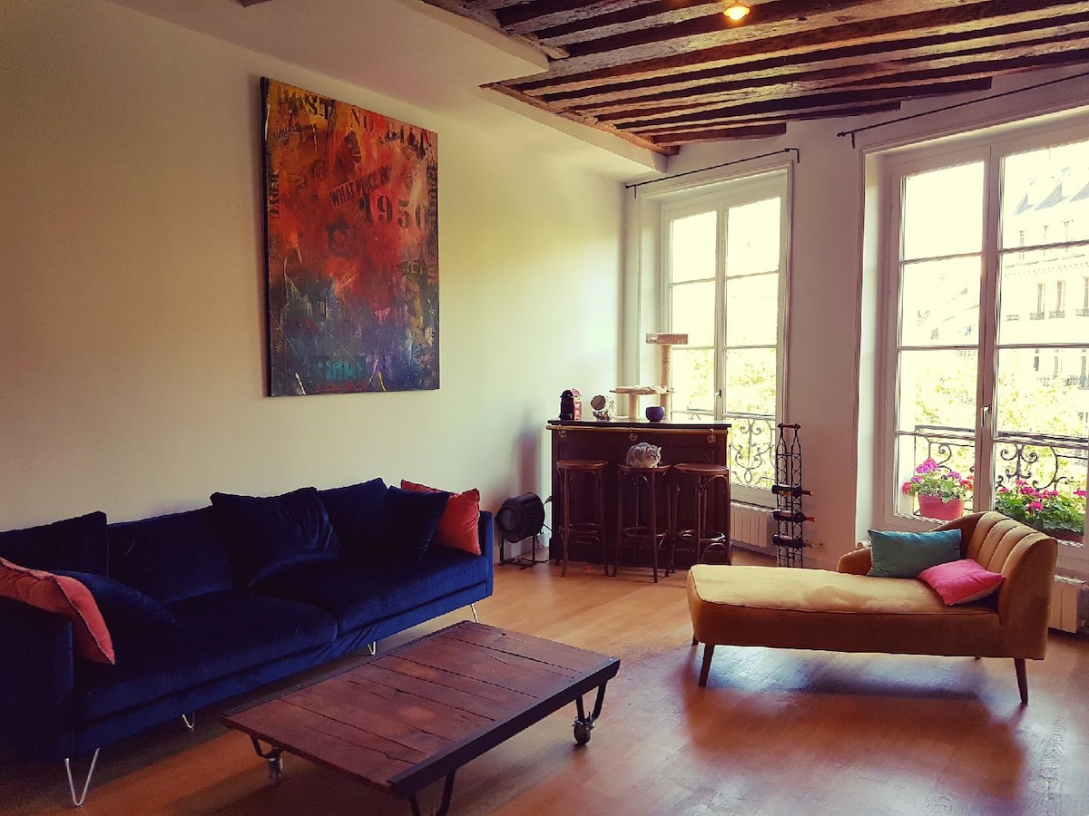 A spacious living area features a deep blue sofa and a vibrant yellow chaise lounge. Large windows allow natural light to fill the room, showcasing a wooden coffee table and a vintage sideboard. Colorful artwork adorns the wall above, adding a touch of character.