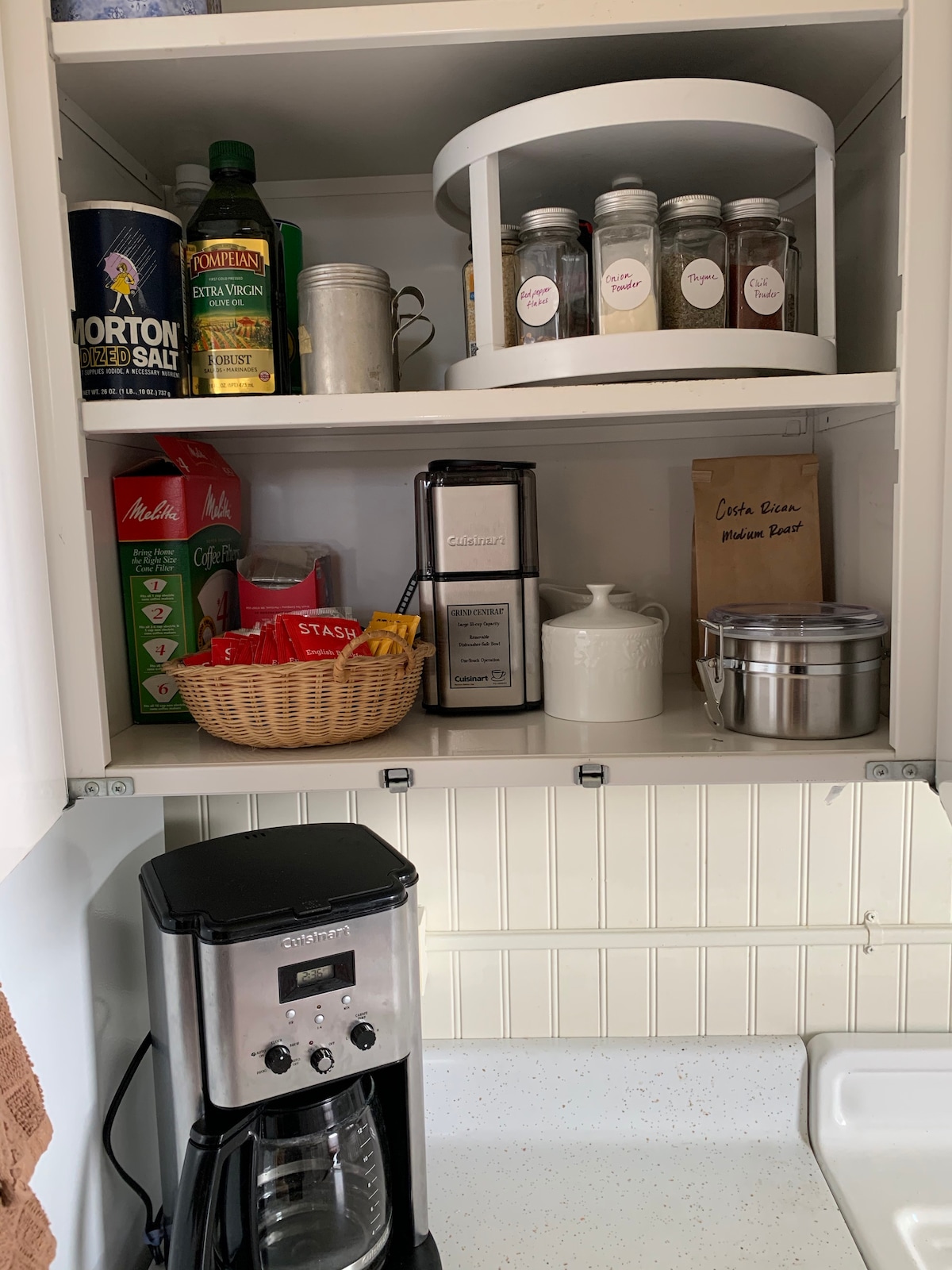 A well-organized kitchen cabinet displays a variety of cooking essentials. Shelves hold spices in labeled containers, a basket with tea bags, and a coffee maker sits on the counter below. An assortment of other kitchen items, including a silver container, is also visible.