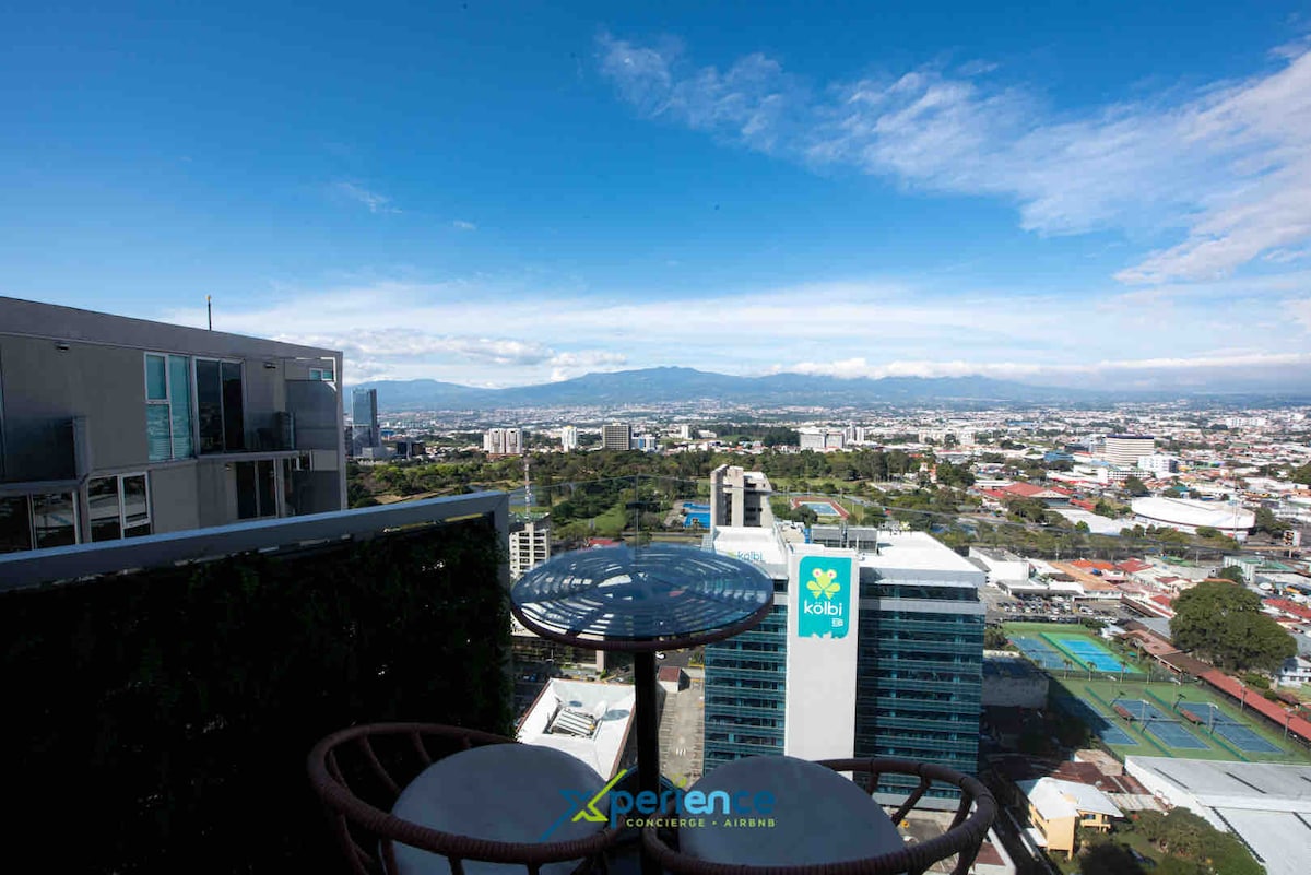 A balcony table with two chairs overlooks a panoramic view of the San José valley, featuring distant mountains and the cityscape. Clear skies enhance the spacious feel of the outdoor area, inviting guests to enjoy the scenery.