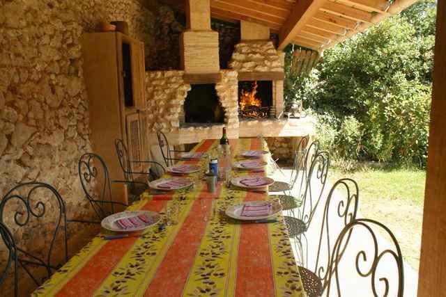 An outdoor dining area features a long table set with colorful plates and surrounded by ornate iron chairs. Two stone barbecue grills can be seen in the background, with a lush green garden providing a natural backdrop. The space is shaded by a wooden roof.