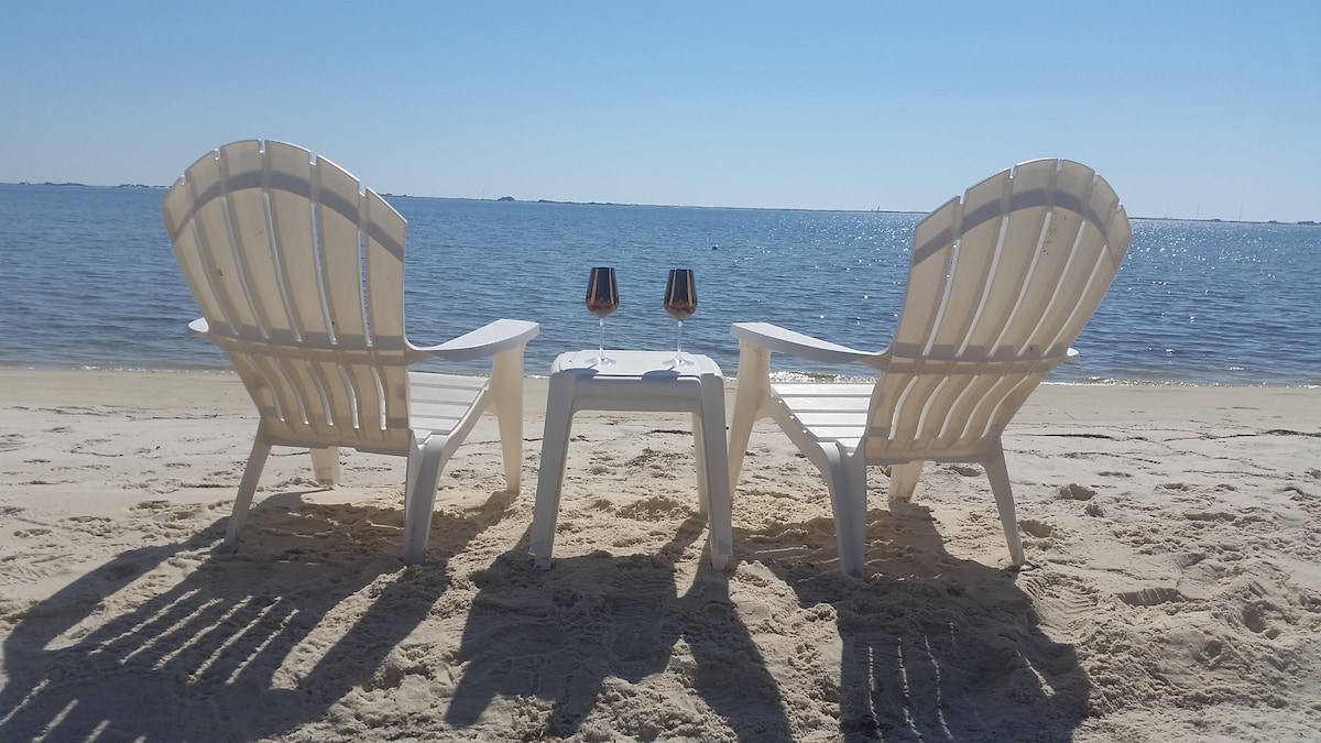 Two white adirondack chairs are positioned on the sandy beach, facing the tranquil water of the Santa Rosa Sound. A small table sits between the chairs, topped with two glasses. The scene is bathed in bright sunlight, with a clear blue sky above.