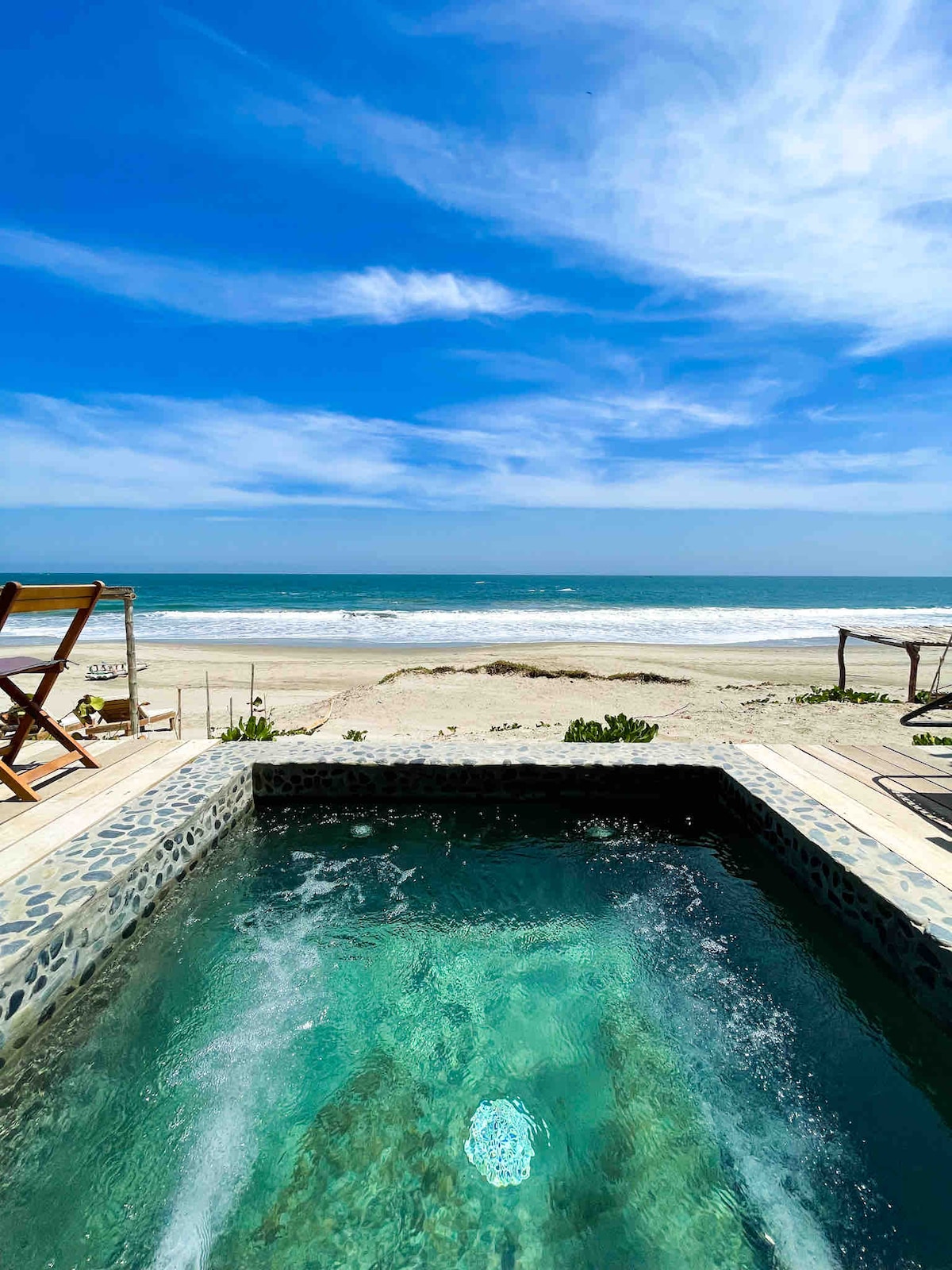 A private terrace features a mini pool with hydromassage jets, surrounded by natural stone and wooden decking. The ocean stretches out in the background, with waves gently breaking on the sandy shore under a bright blue sky.