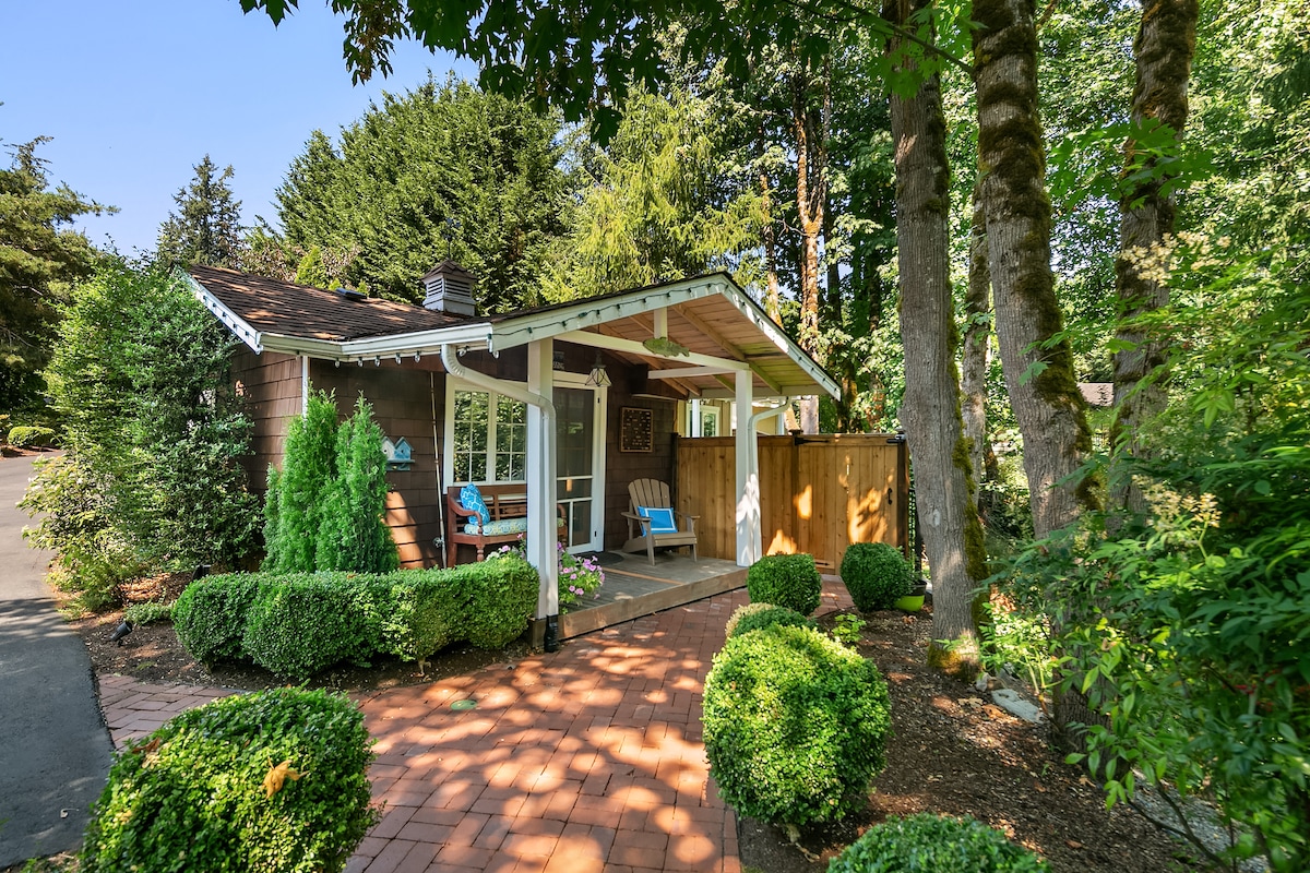 A charming cottage exterior is framed by lush greenery and landscaped shrubs. A paved pathway leads to the entrance, where a covered porch is furnished with two chairs. The surrounding trees provide a sense of privacy and tranquility.