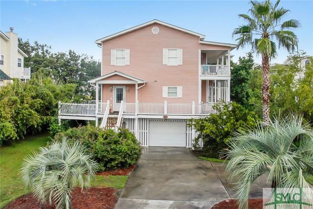 A two-story pink house is set amidst lush greenery and palm trees. The elevated structure features a spacious front porch with white railings, leading down to a concrete driveway with a garage underneath. The surrounding landscape is neatly landscaped with mulch and vibrant plants.