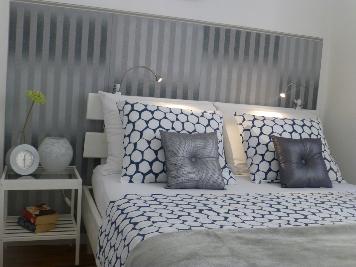 A serene bedroom is presented, featuring a spacious bed with geometric-patterned bedding. Two matching pillows enhance the decor, while bedside lamps provide soft lighting. A small nightstand holds a clock and decorative items, contributing to an organized and elegant environment.