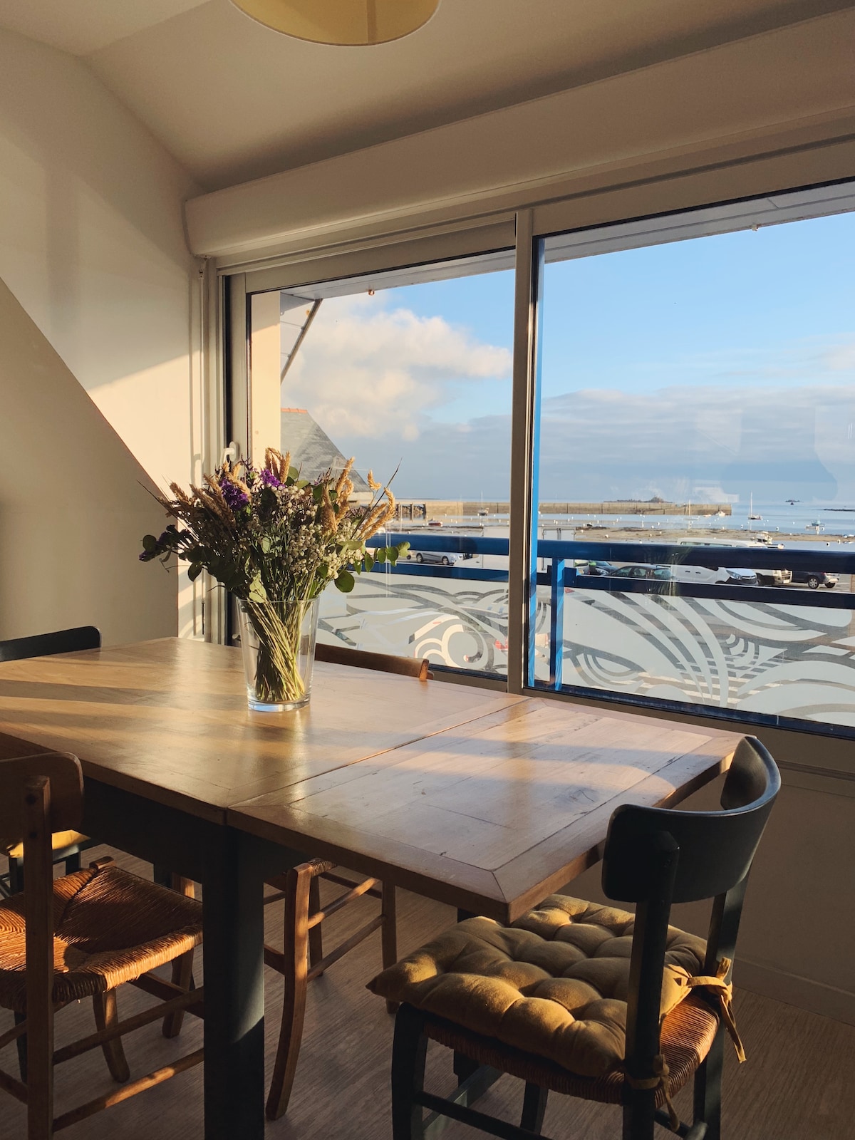 A dining area features a large wooden table surrounded by several chairs. A flower vase sits in the center, while large windows offer a view of the sea and harbor. Natural light illuminates the space, creating a bright and airy environment.
