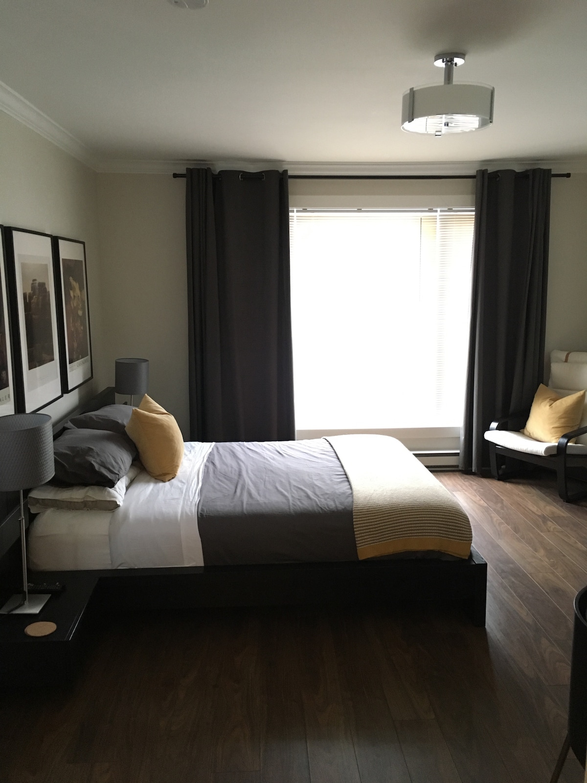 A well-appointed bedroom features a spacious bed with neutral bedding and a decorative yellow accent pillow. Two nightstands with lamps flank the bed, and a chair is positioned beside a large window dressed with dark curtains, allowing natural light to fill the space.