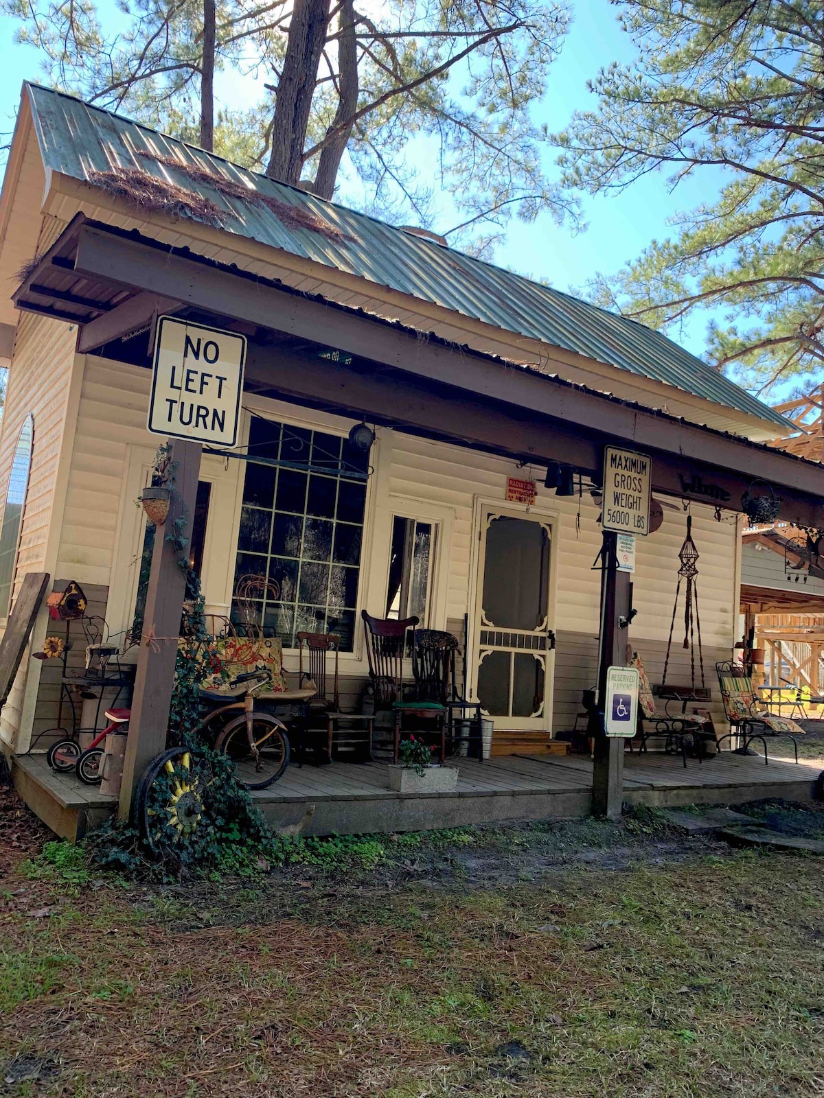 A charming exterior of a country shack is visible, featuring a covered porch with various seating options, including rocking chairs. The building is surrounded by tall pine trees, with a mix of rustic signage and decorative elements adding character to the inviting entrance.