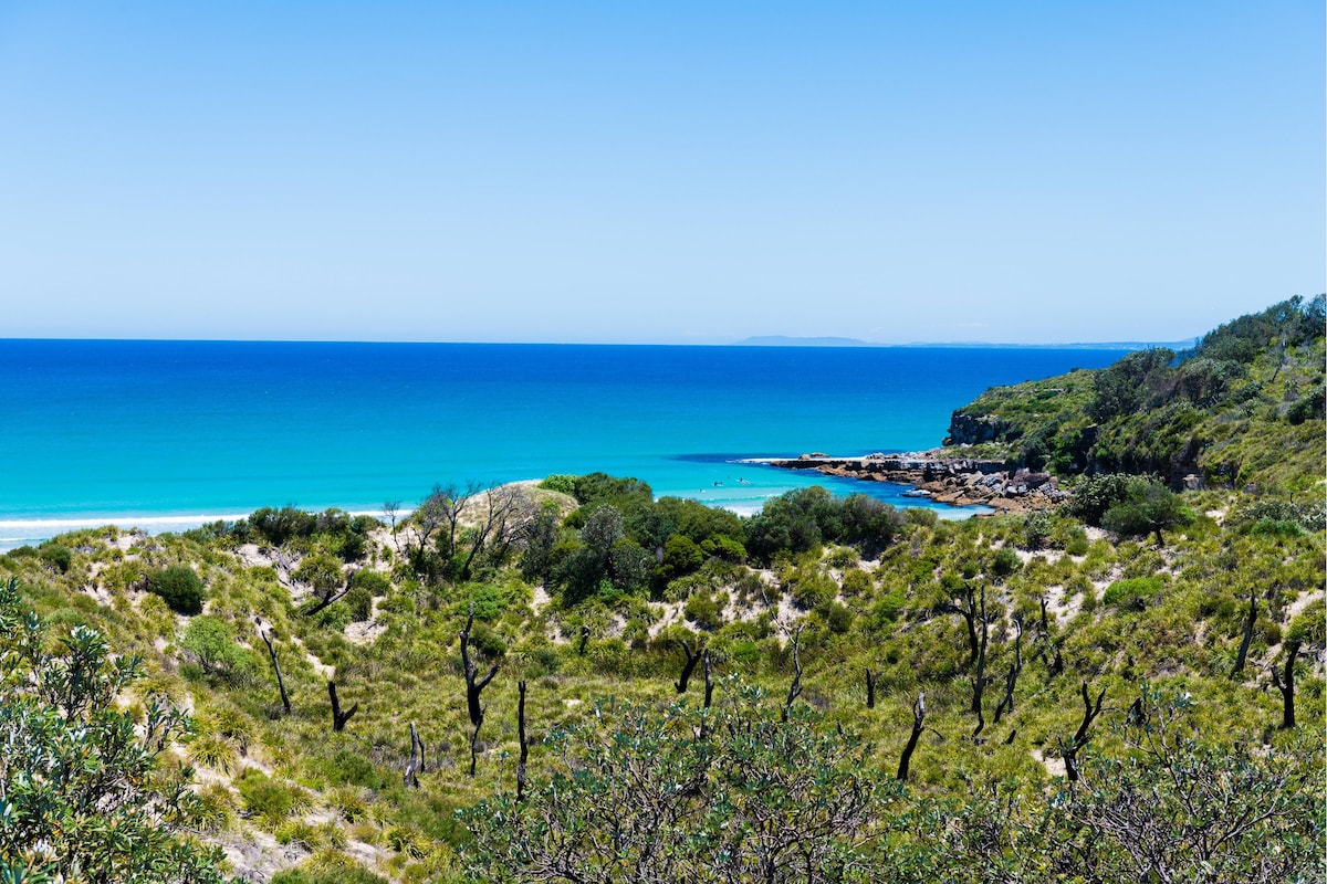 A scenic coastal landscape is presented, featuring a calm turquoise sea that meets the sandy shore. Lush greenery and trees frame the view, leading towards a small inlet. The clear blue sky complements the tranquil atmosphere, creating a peaceful retreat by the water.