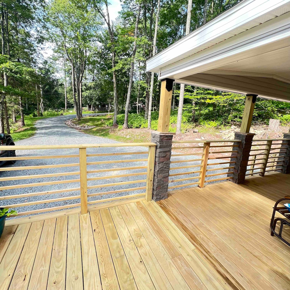 A spacious wooden deck overlooks a gravel path leading into a wooded area. Surrounding trees and greenery create a natural setting, while the railings provide safety and a welcoming touch. The scene offers a glimpse of the tranquil outdoor environment offered by the property.