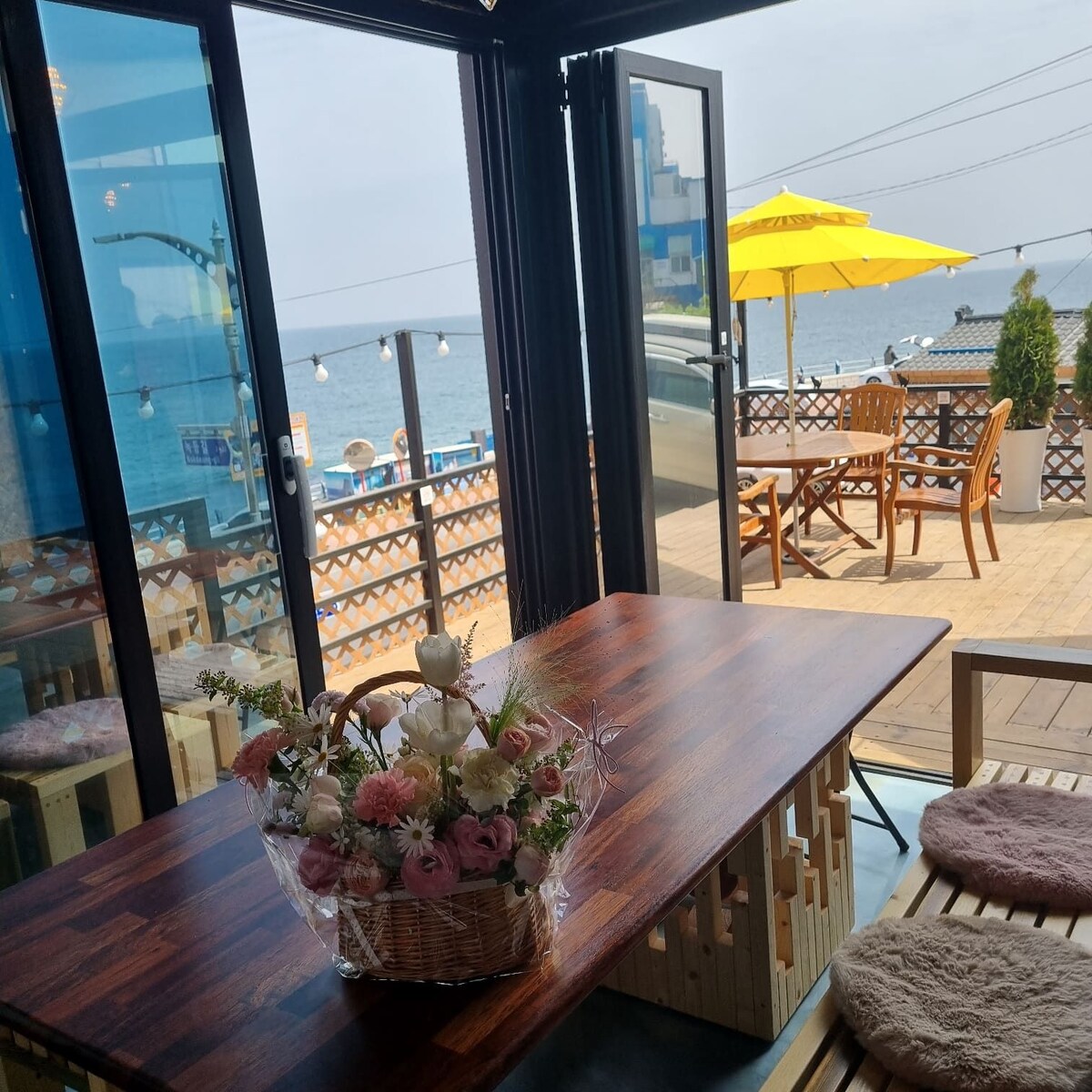 A spacious dining area is seen through large glass doors, offering a view of the ocean. A wooden table is set with a floral arrangement, and outdoor seating is available on the deck, shaded by a yellow umbrella.