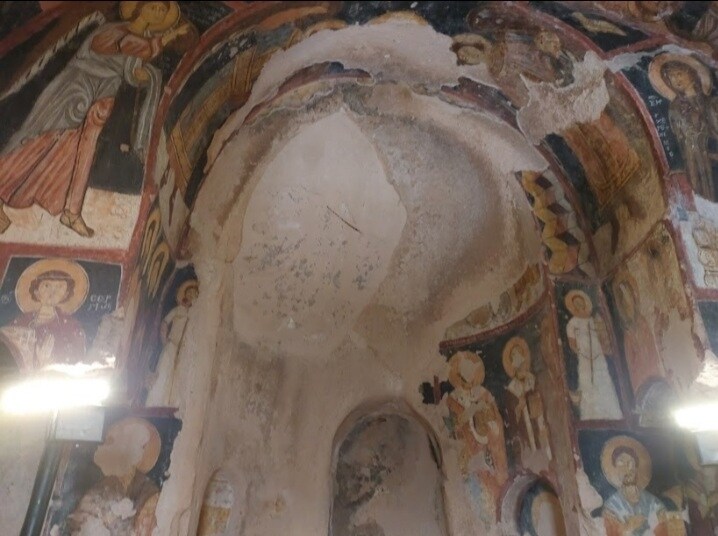 The interior of a historical site is depicted, featuring ancient frescoes on the walls. Religious figures are illustrated in vibrant colors on the walls, with remnants of intricate arches visible overhead. Soft light emanates from hidden sources, highlighting the texture of the aged surfaces.