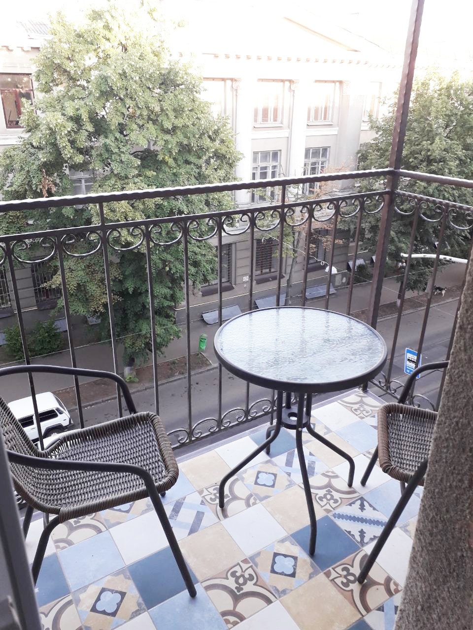 A cozy balcony is equipped with a round glass table and two woven chairs. The patterned tiled floor complements the view of trees and a street below. Railings frame the space, allowing for an inviting outdoor setting.