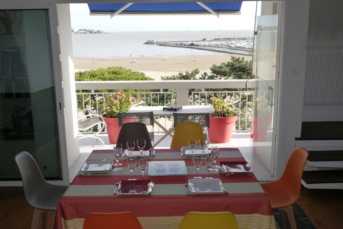 A dining area features a table set with glassware and colorful chairs, overlooking a beach view through large glass doors. Planters with greenery frame the scene, highlighting the outdoor space, while the calm coastline and harbor are visible in the background.