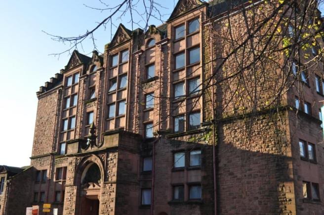 The exterior of a converted school building is shown, featuring a textured stone façade with prominent architectural details. Large windows punctuate the structure, allowing natural light to enter. The entrance is framed by a decorative arch, surrounded by a tree's branches.