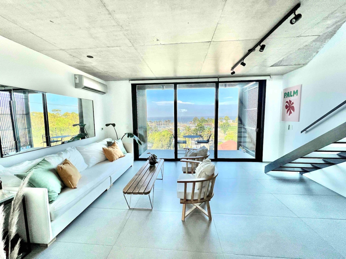 A spacious living area is highlighted by large glass doors, offering views of the surrounding landscape. A comfortable white sofa is complemented by a wooden coffee table and two light-colored chairs. Natural light fills the room, enhancing the modern and minimalist design.
