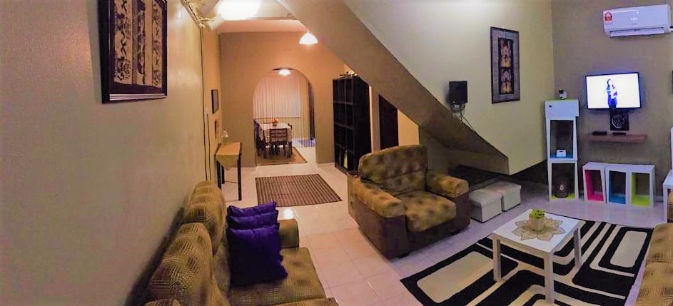 An open living area is furnished with two cozy sofas, a coffee table, and decorative cushions. A flat-screen TV is mounted on the wall, with a dining area visible in the background. Natural light brightens the space, enhancing the inviting atmosphere.