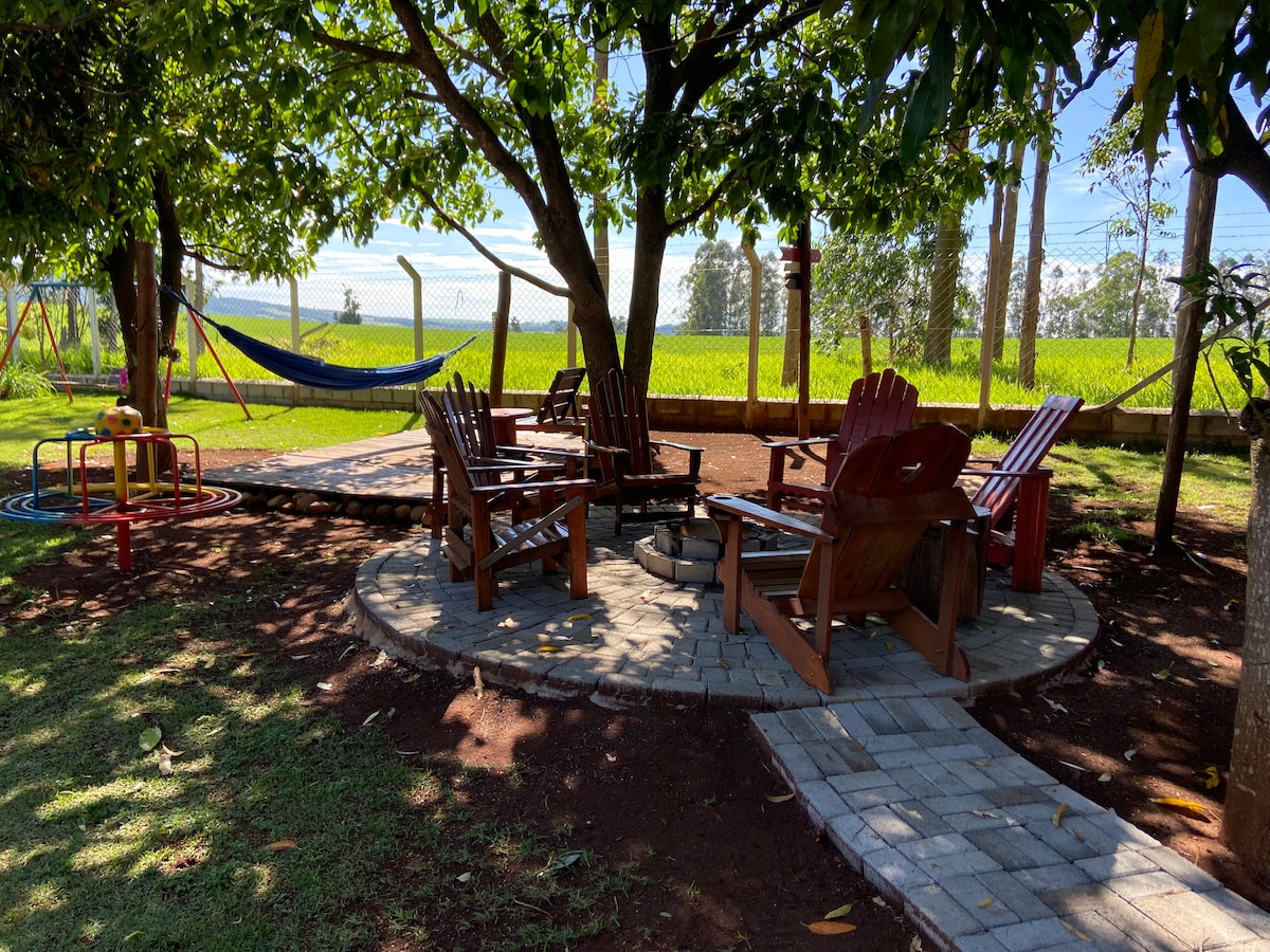 A shaded outdoor seating area features rustic wooden chairs arranged around a stone fire pit. Nearby, a hammock is gently swaying among the trees, and a small playground structure is visible in the background. Lush greenery complements the serene environment.