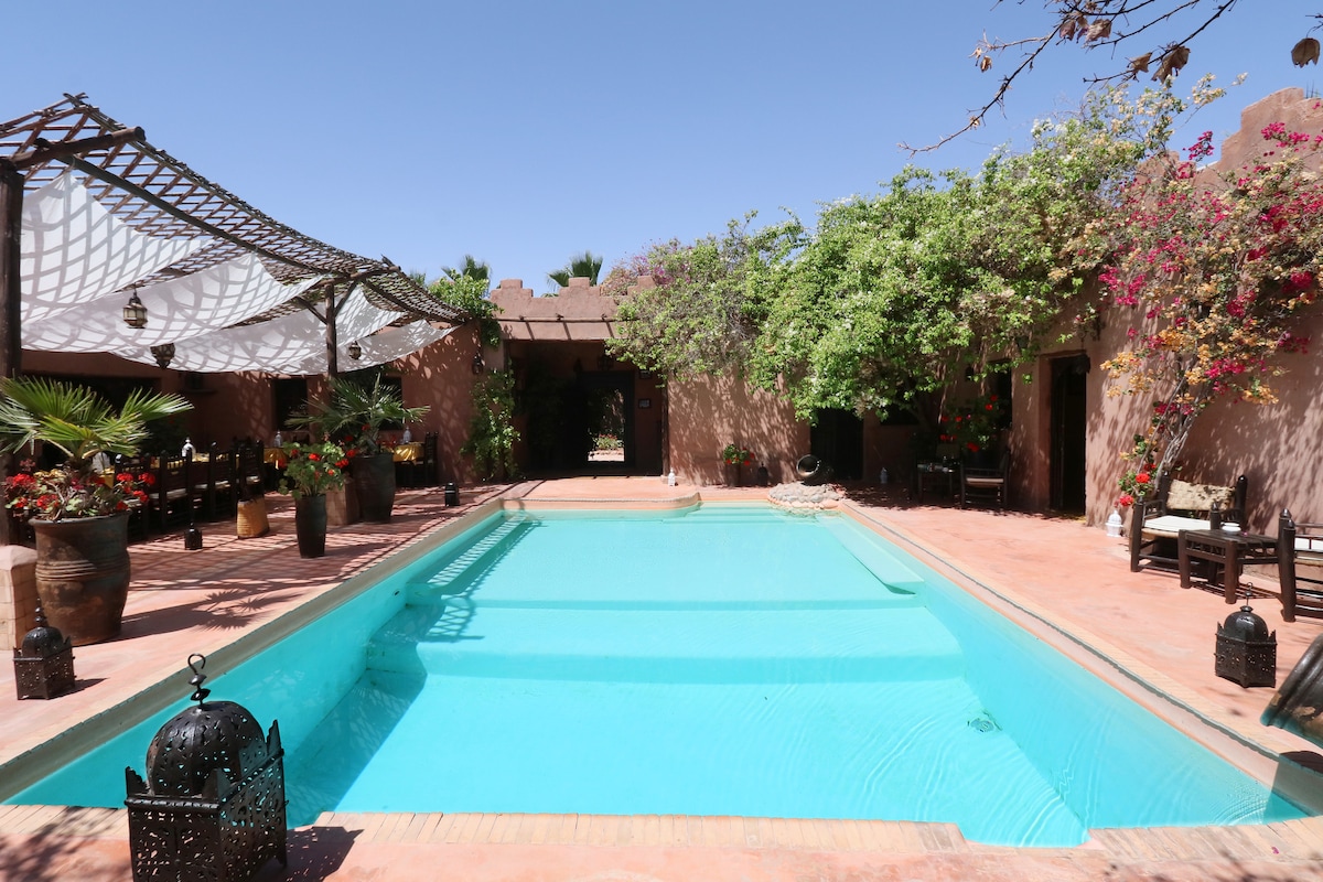 A serene courtyard is showcased, featuring a central swimming pool surrounded by lush greenery and vibrant flowers. Rustic seating areas are positioned around the pool, creating an inviting space for relaxation. Traditional lanterns are placed throughout the area, enhancing the tranquil atmosphere.