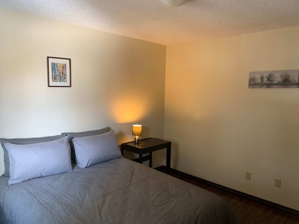 A calm bedroom features a spacious bed dressed in gray linens, accompanied by two pillows. A bedside table with a lamp illuminates the space. A piece of artwork hangs on the wall, adding a subtle touch to the neutral-colored room.