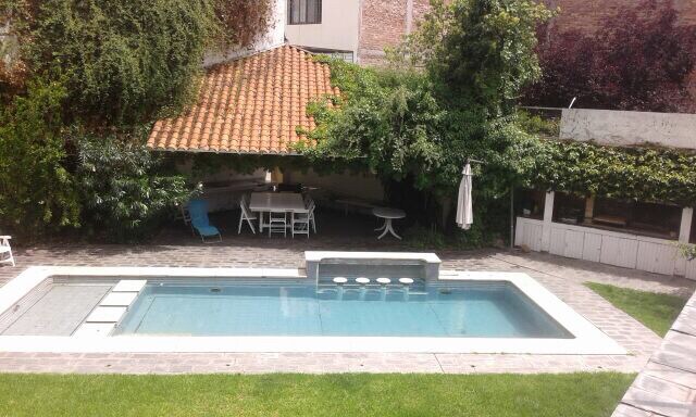 A spacious pool is framed by a well-maintained patio and greenery. A shaded area with seating is positioned nearby, featuring a large table and chairs. The surrounding grass is neatly trimmed, providing a relaxing outdoor space.