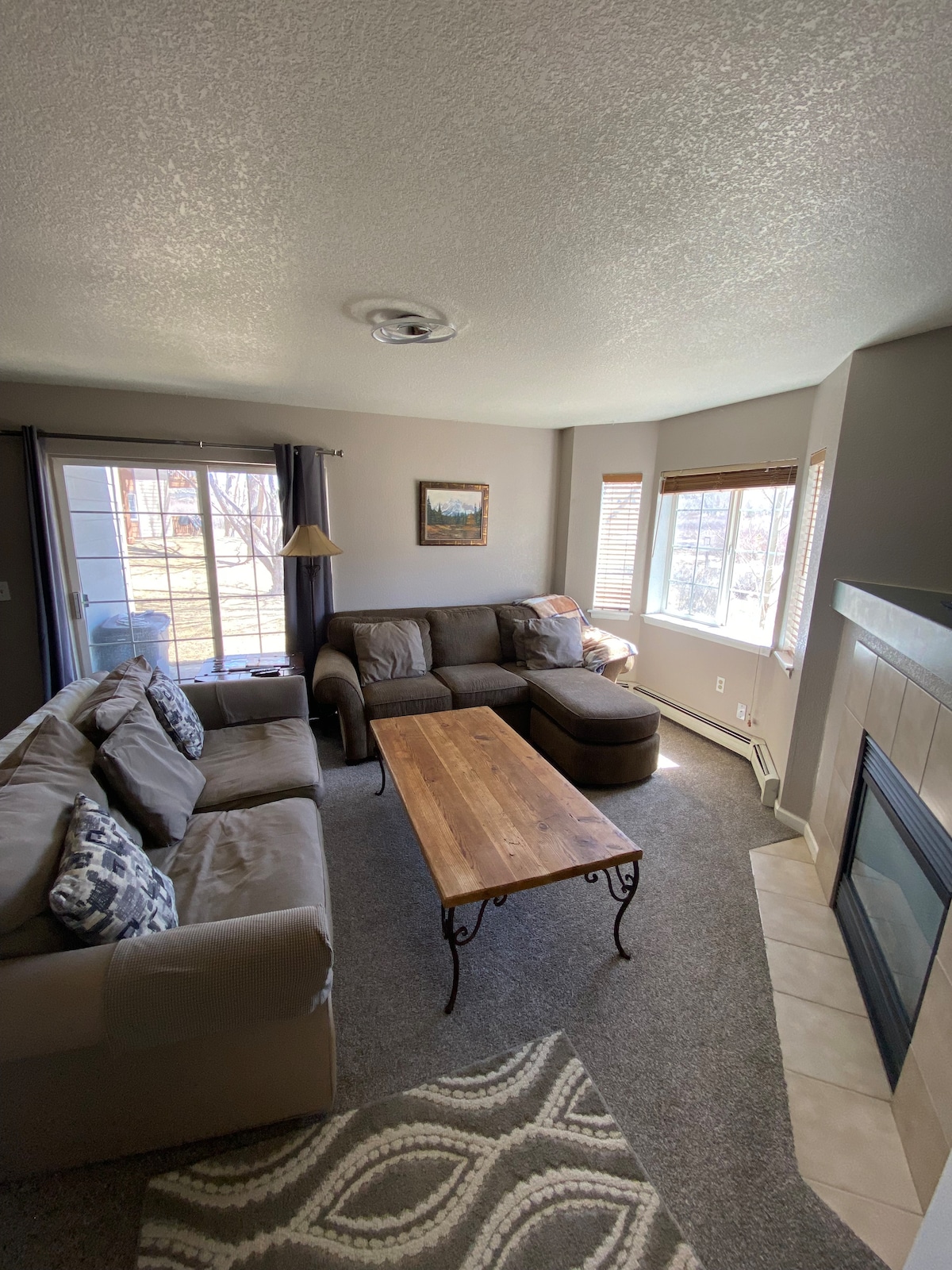 A cozy living area is showcased, featuring a large sectional sofa and a wooden coffee table. Natural light enters through large windows, highlighting the neutral color palette. A stone fireplace adds a touch of warmth, while subtle decorative accents are placed on the sofa.