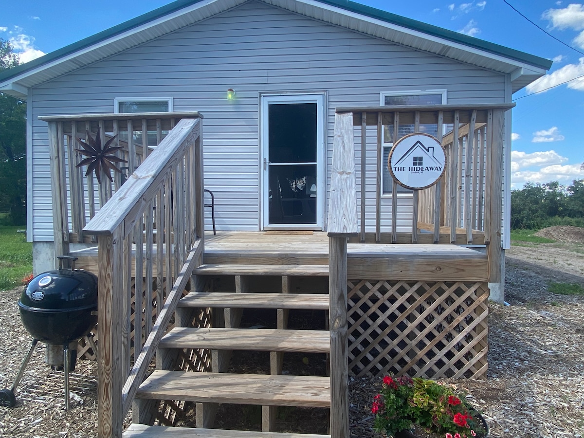The exterior of the cozy industrial home is featured, showing a wooden deck with steps leading up to the entrance. A charcoal grill is positioned beside the steps, while flowers are planted near the base of the deck. The house is surrounded by greenery.