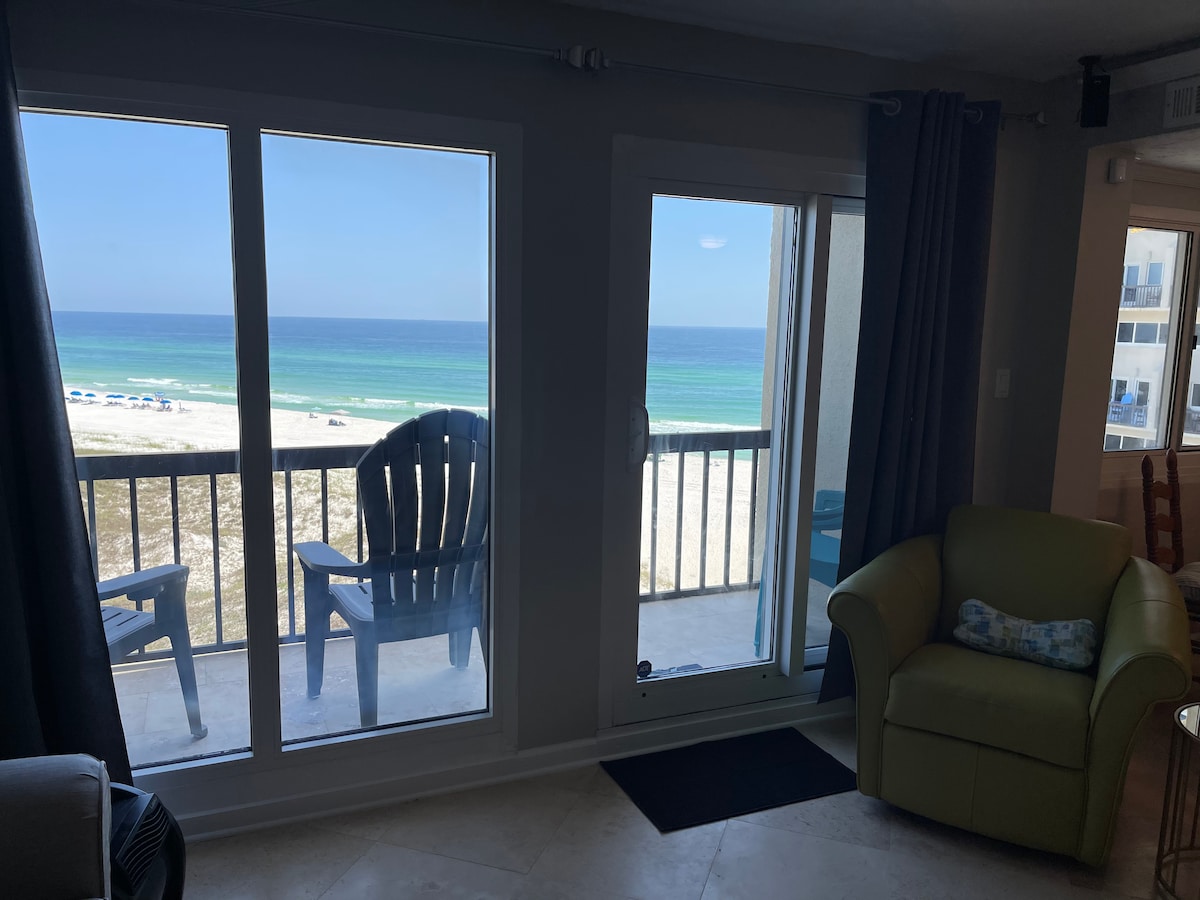 Views of the Gulf are captured through expansive glass doors. A pair of outdoor chairs is visible on the balcony, providing a place to relax. Natural light brightens the room, highlighting a green armchair positioned nearby.