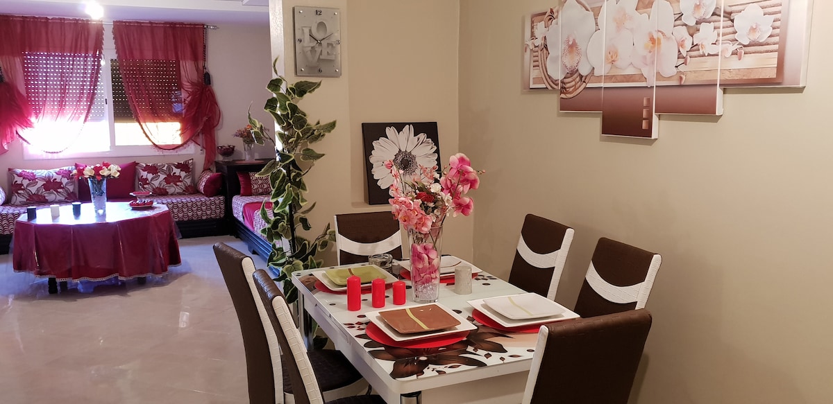 A dining area featuring a white table set for six, accented by tasteful decorative elements. Fresh flowers are arranged in a vase at the center. The spacious living area is visible in the background, with comfortable seating and bright curtains complementing the decor.