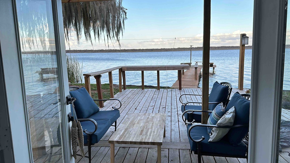 A wooden deck offers a serene lake view, featuring two cushioned chairs facing a small table. Gentle ripples are visible on the water, while a boat dock can be seen in the background, surrounded by lush greenery.
