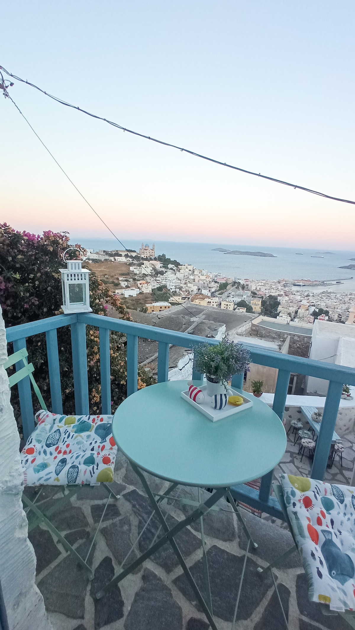 A charming balcony features a round table with two decorative chairs, surrounded by vibrant cushions. A small potted plant sits at the center, while the scenic view of the town and sea creates a calming backdrop at dusk.