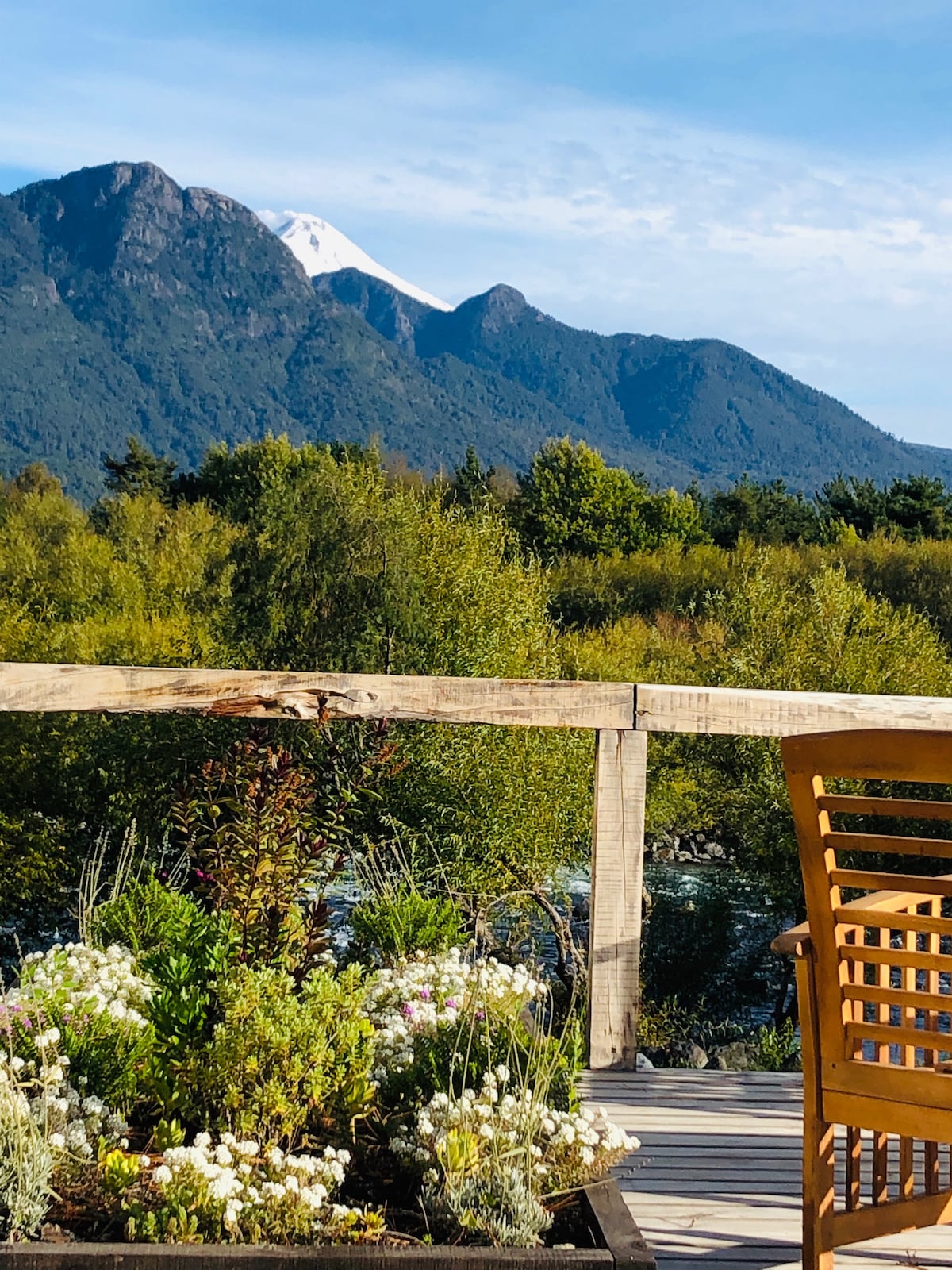 A wooden deck overlooks a lush landscape with flowering plants in the foreground. A mountain range is visible in the background, displaying a snow-capped peak against a clear blue sky, while greenery frames the scene, suggesting tranquility and natural beauty.