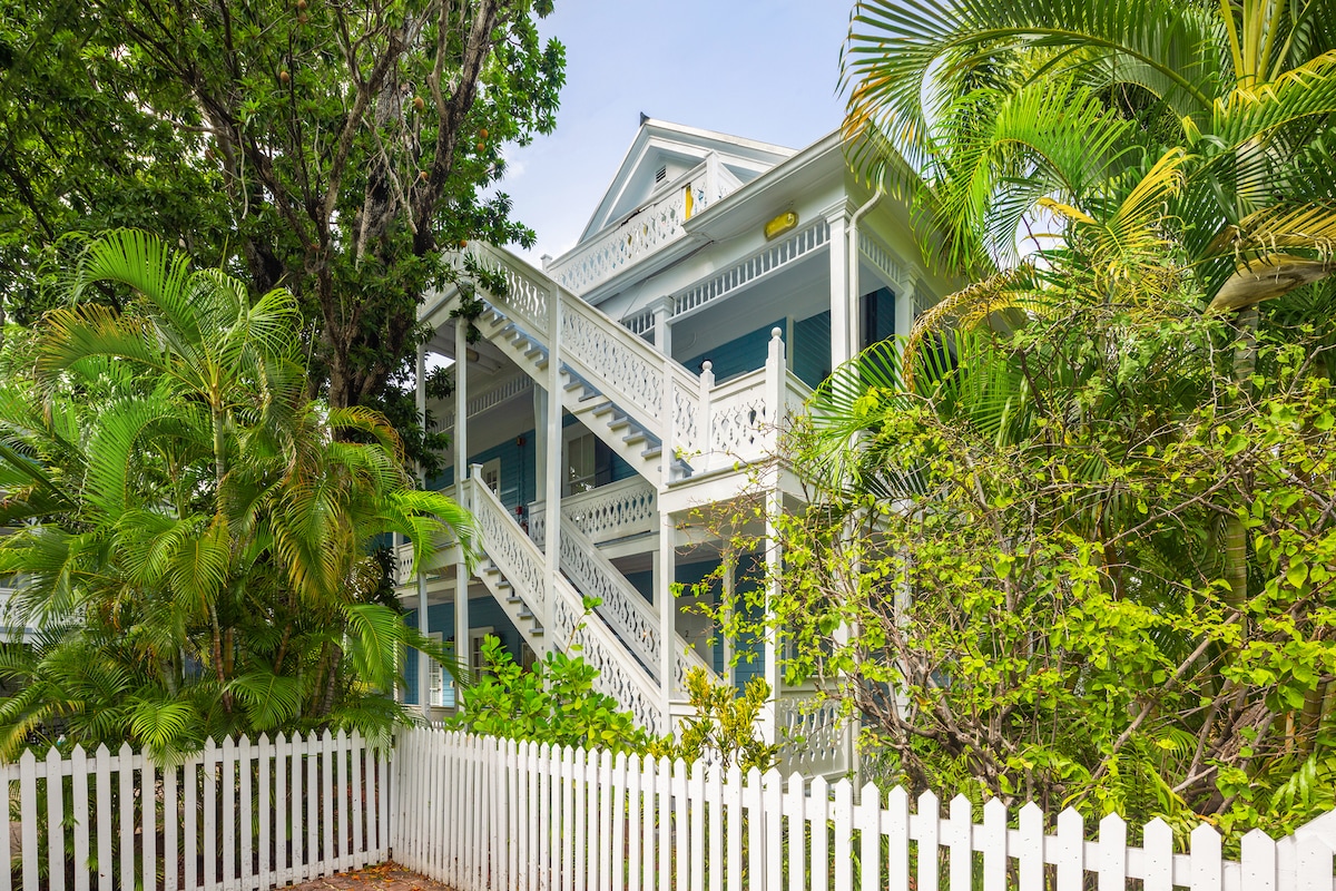 A tropical setting frames the exterior of a multi-level building, showcasing a charming architectural design with balconies and a decorative staircase. Lush greenery surrounds the structure, providing a sense of tranquility and seclusion against a clear sky.