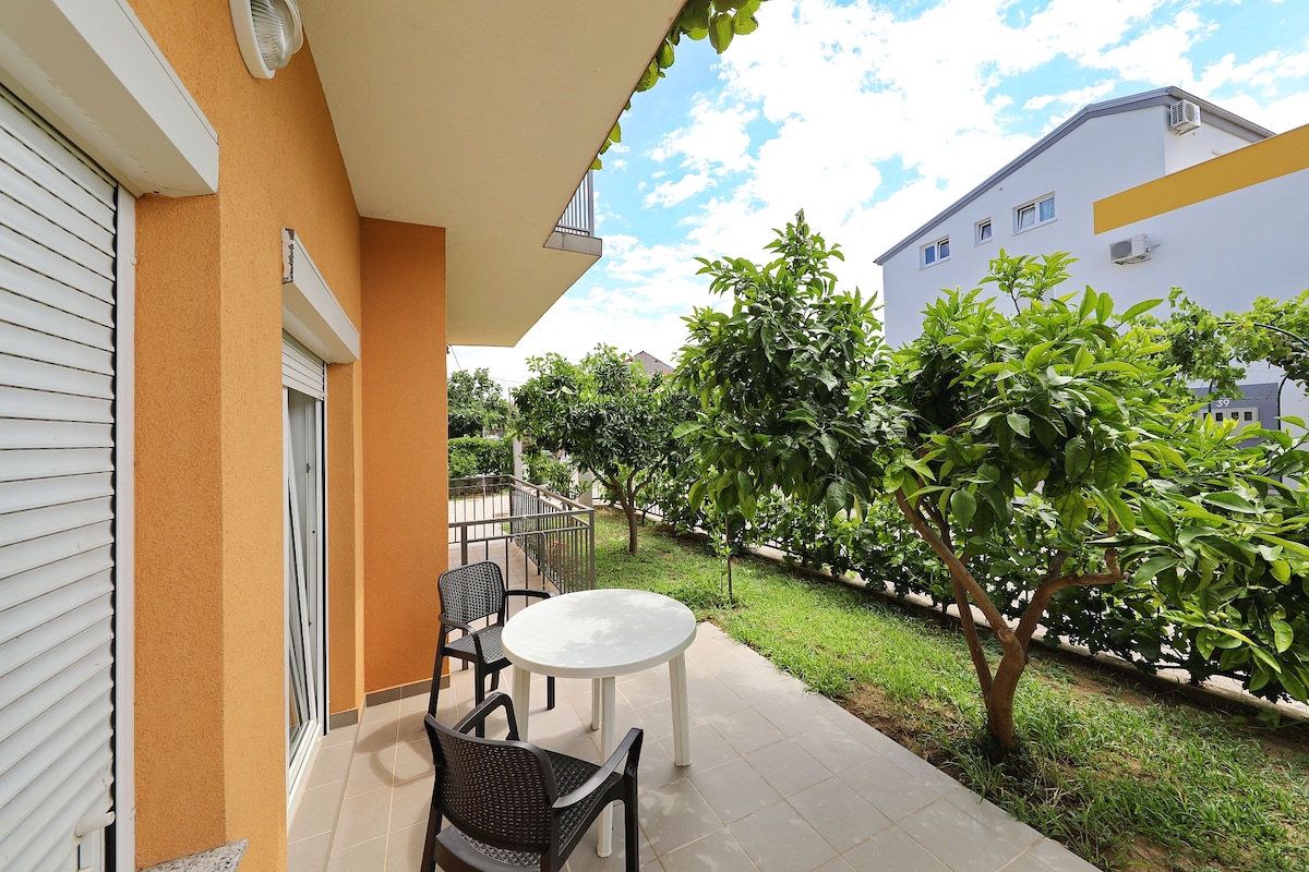 An outdoor terrace features a round table surrounded by two chairs. Lush greenery fills the space, bordered by a low fence. The building exterior is painted in warm tones, while the sky above is partly cloudy, allowing natural light to illuminate the area.