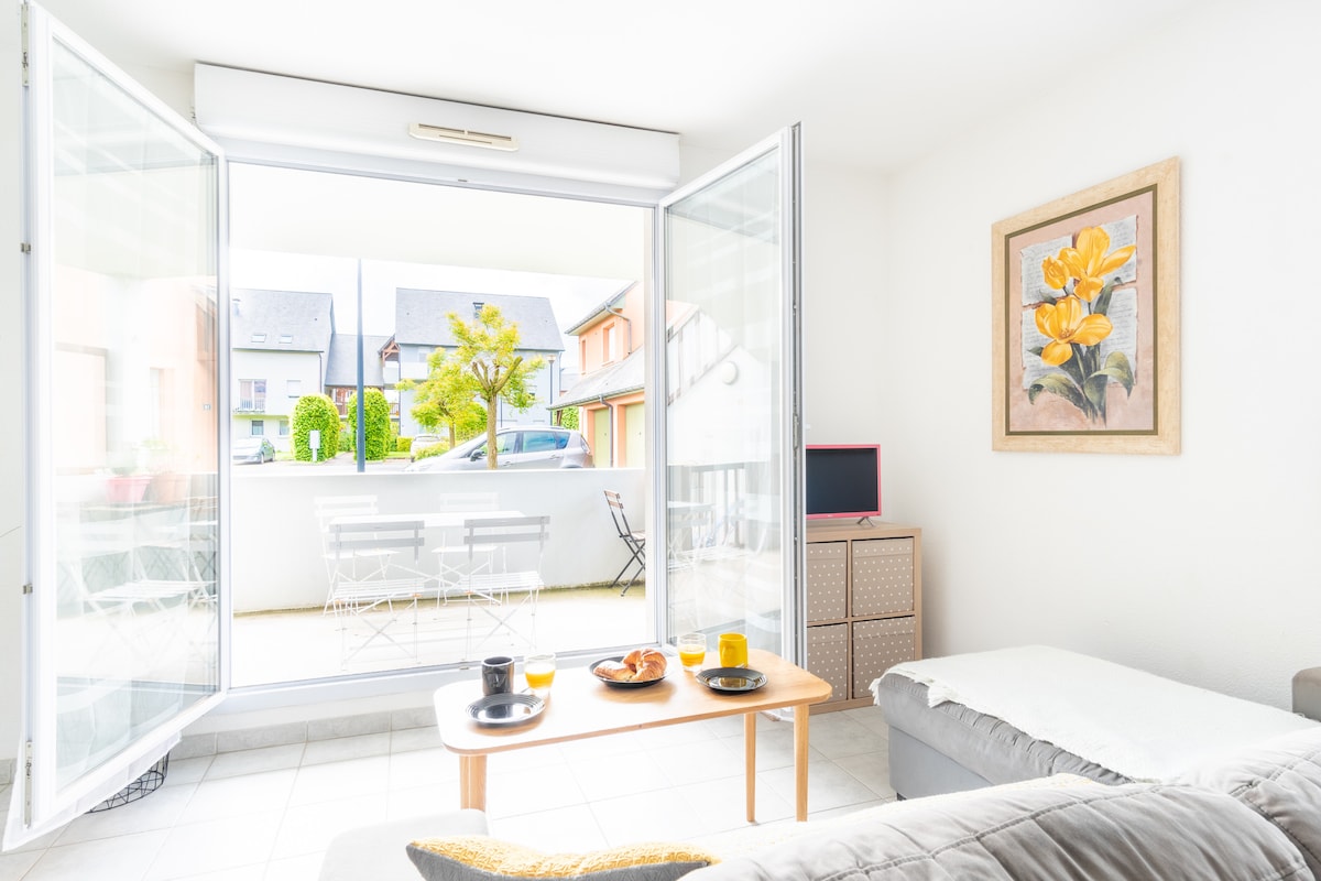 A bright and airy living area is presented with large glass doors that open to a balcony. A small coffee table is set with refreshments, while a comfortable sofa is positioned nearby. A piece of floral artwork adorns the wall, and a television is visible in the corner.