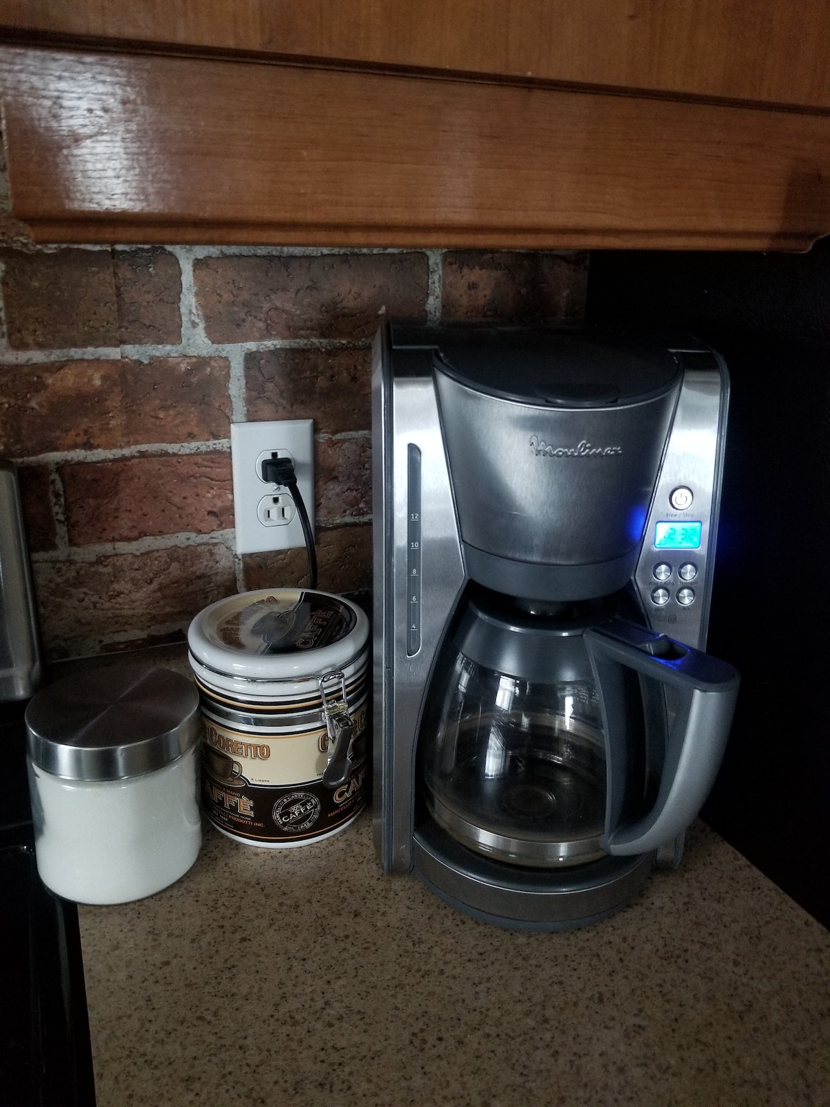 A coffee maker is positioned on a countertop beside a container of coffee grounds and a sugar jar. The backdrop features exposed brick, adding a rustic touch to the kitchen space.