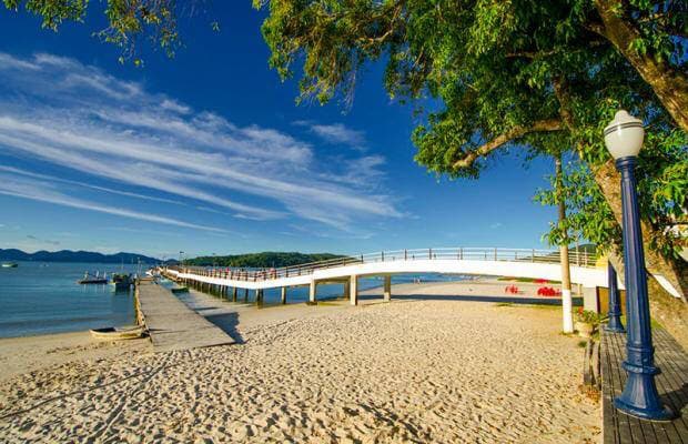 A wide sandy beach is framed by lush green trees and a clear blue sky. A wooden boardwalk stretches out over the water, leading toward the horizon. A lamppost stands along the pathway, providing a sense of connection to the scenic landscape.