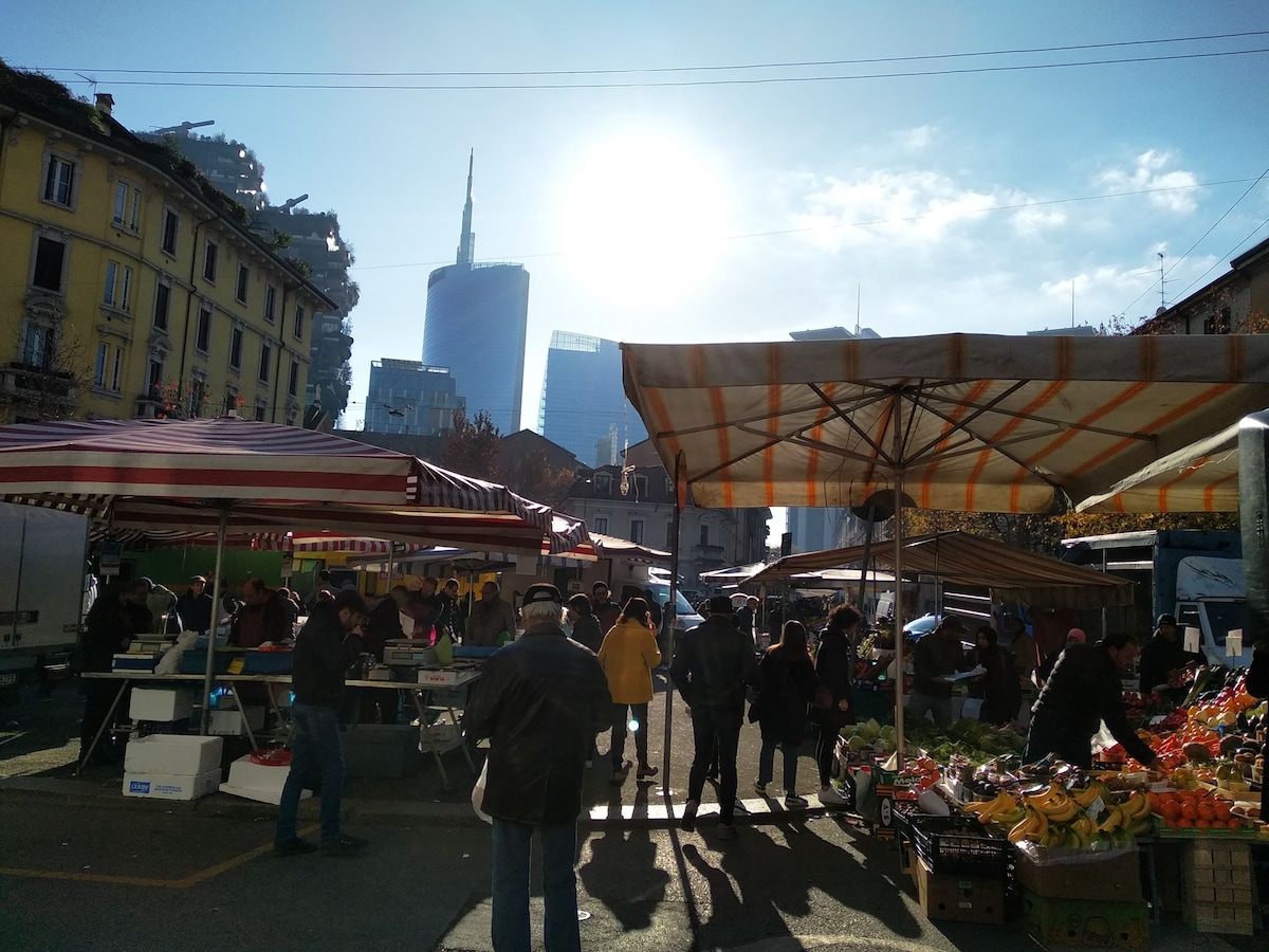 A bustling outdoor market is featured, with striped canopies providing shade over vendor stalls filled with fresh produce. Shimmering skyscrapers are visible in the background, framed by a bright blue sky. Groups of people can be seen engaging in shopping and socializing.
