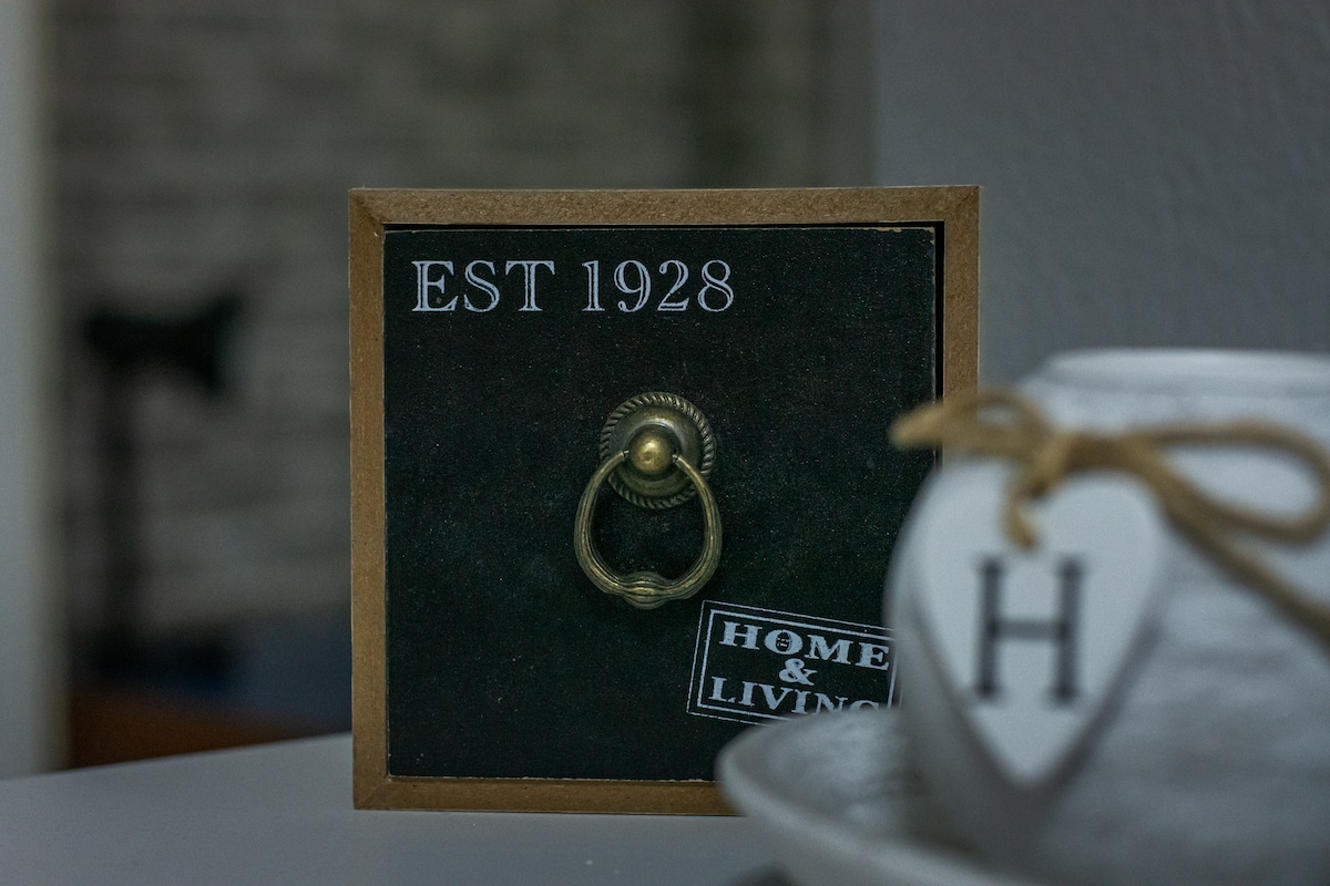 A decorative wall plaque is displayed, featuring the text 'EST 1928' and 'HOME & LIVING'. An antique-style ring pull handle is positioned at the center, enhancing the vintage feel. A simple ceramic pot with a heart label and the letter 'H' can be seen nearby.