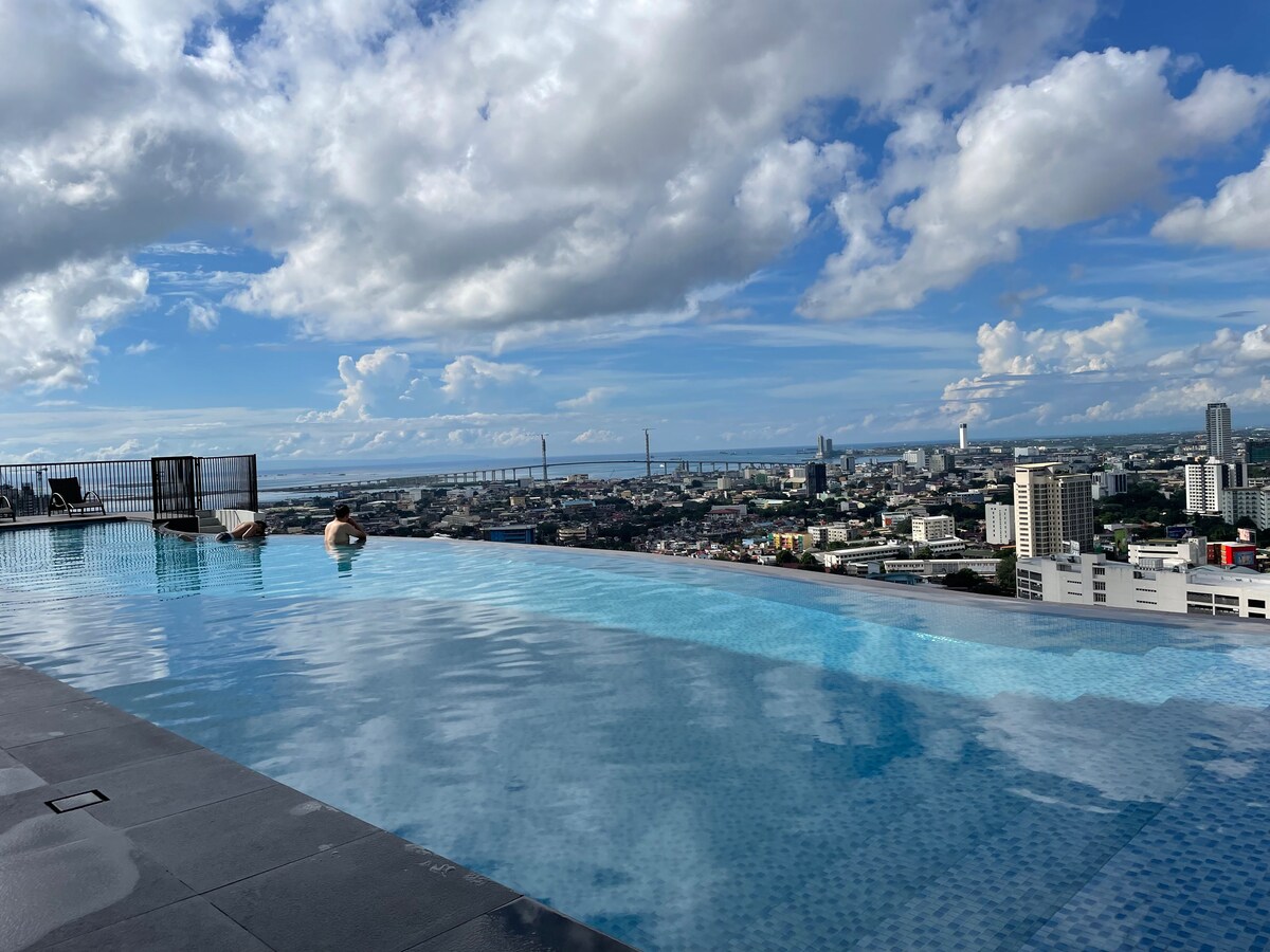 An infinity swimming pool extends along the rooftop, offering stunning views of the city and Mactan channel. The calm water reflects the sky, while guests are seen enjoying the serene environment. The landscape is framed with buildings and greenery in the distance.