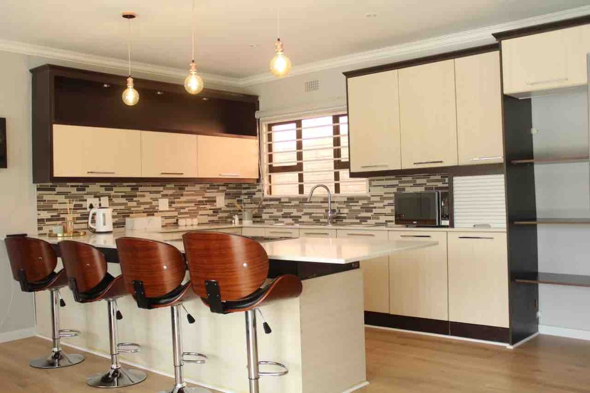 A modern kitchen is displayed with sleek cabinetry in light and dark tones. A spacious island is complemented by four high-backed stools. Neutral tile work adds texture to the backsplash, while warm lighting enhances the overall ambiance.