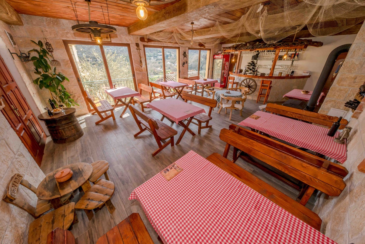 An inviting dining area features wooden tables with red checkered tablecloths, surrounded by a rustic décor. Large windows provide natural light and views of the surrounding landscape. Bar stools are positioned at a counter in the background, enhancing the communal dining experience.