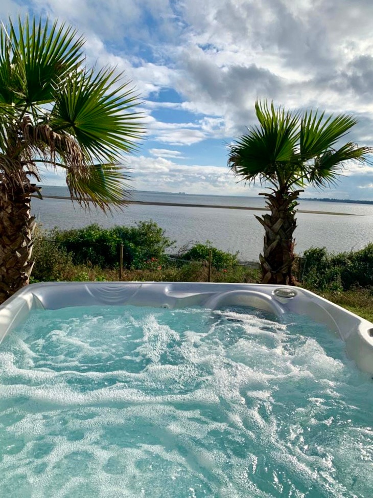 A hot tub is situated on a deck, surrounded by palm trees that frame an expansive view of calm waters and a cloudy sky. Bubbles can be seen rising in the water, creating a soothing atmosphere perfect for relaxation.