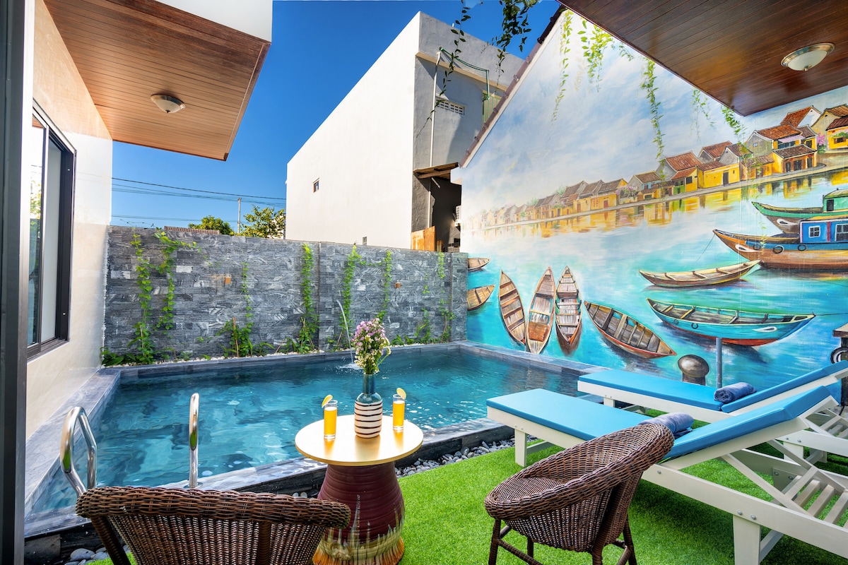 A private swimming pool is surrounded by a lush green lawn, featuring two lounge chairs positioned nearby. A table is set with two candles, adding to the tranquil atmosphere. A mural of boats and a waterfront scene decorates the wall, enhancing the serene setting.
