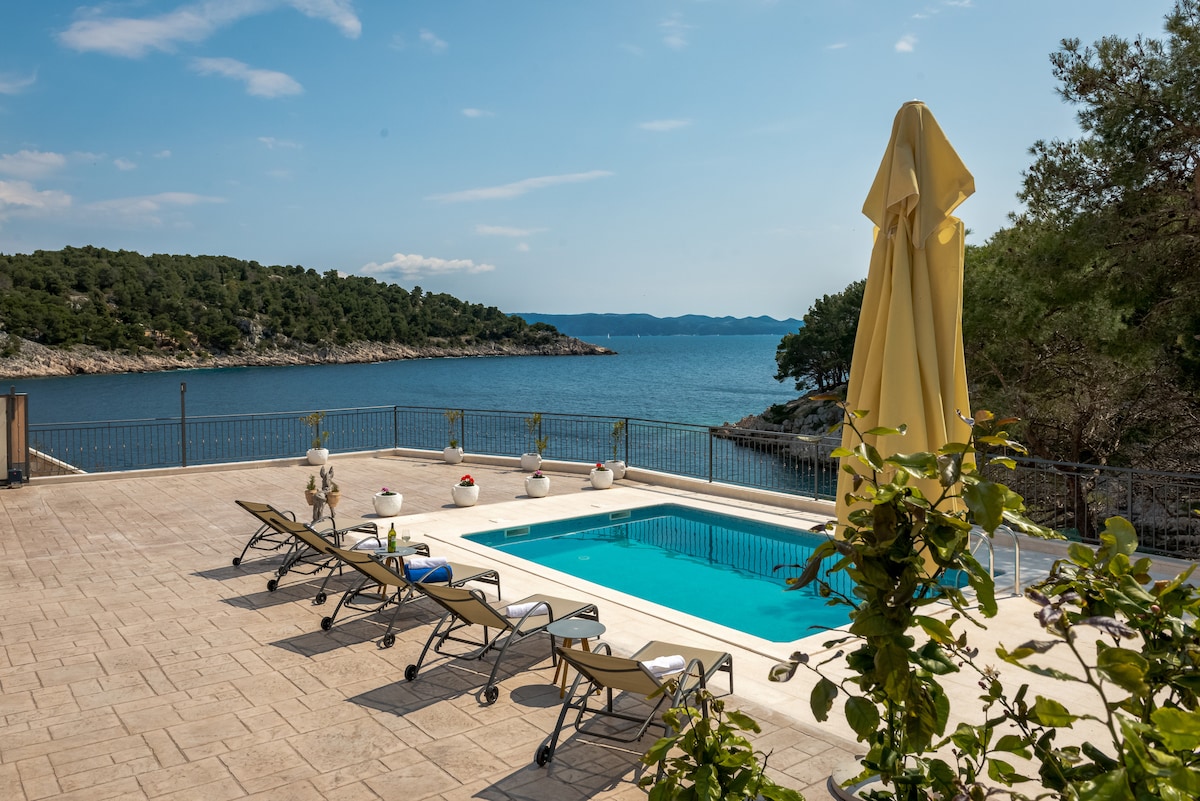 An outdoor terrace overlooks the sea, featuring a private swimming pool surrounded by lounge chairs. A large umbrella provides shade, and decorative planters add a touch of greenery. The expansive view of the bay and rocky shoreline enhances the serene atmosphere.